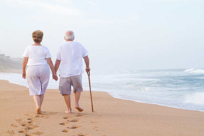 An older couple walks barefoot on the beach, the man uses a cane.