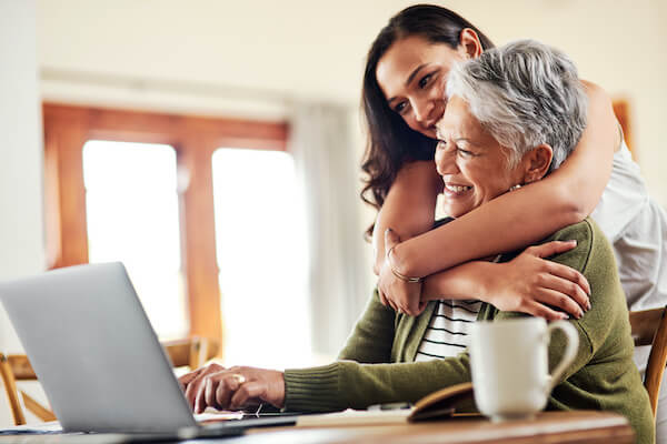 A smiling older woman sits at a laptop in a sunny room, with her adult daughter hugging her.